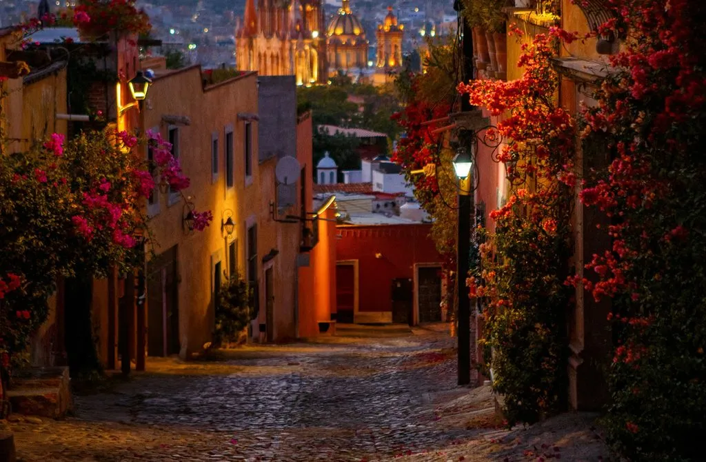 A quiet cobblestone side street in San Miguel de Allende lined with bougainvillea at dusk, the Parroquia lit in the distance